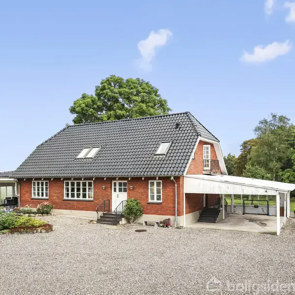 Et rødt murstenshus med sort tegltag står på en grusbelagt gårdsplads. Til venstre ses en lille have med træer; til højre holder en hvid bil under en carport.