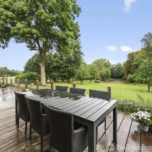 Terrasse med sort spisebord og seks stole står på trædæk. I baggrunden ses en grøn græsplæne med træer og buske, under blå himmel med få hvide skyer.