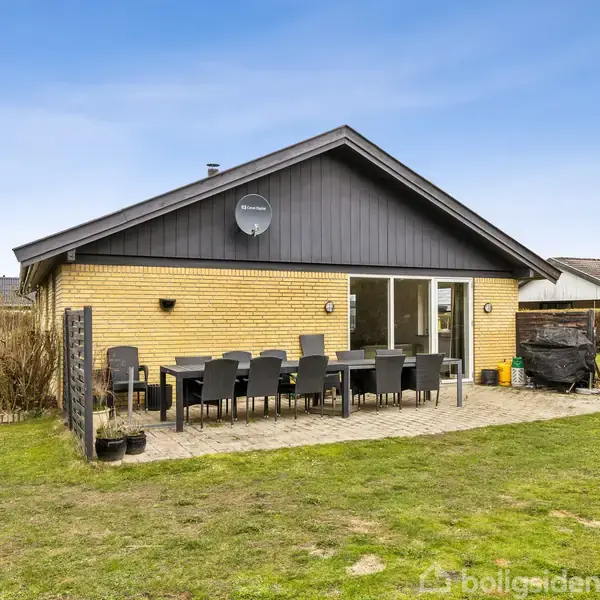 En gul murstenshus har en flad tagkonstruktion. Foran huset står et bord med otte sorte stole på en terrasse. Græsplæne og buske omkranser området.