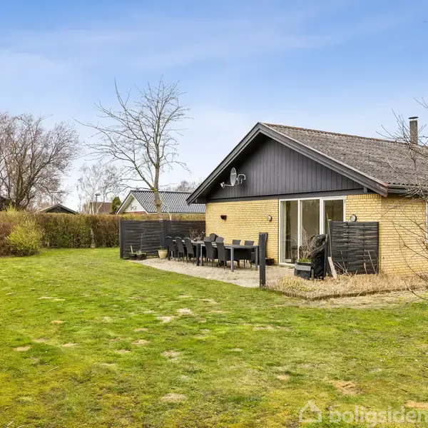 Et gult murstenshus med træbeklædning har en terrasse med havemøbler. Huset er omgivet af en stor græsplæne og nogle træer, placeringen fremstår rolig og naturlig.