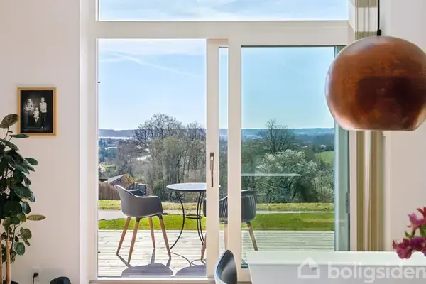 Et rundt bord med to stole står på en terrasse, der har udsigt til en grøn, kuperet landskab under en blå, skyfri himmel. En stor lampe og et indendørs plante ses i forgrunden.