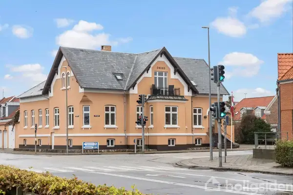 Et stort, beige hus, dateret 1909, står ved et vejkryds med trafiklys. Huset har mørkt tag og hvide vinduer. En blå vejskilt er placeret foran huset, der er omgivet af fortove.