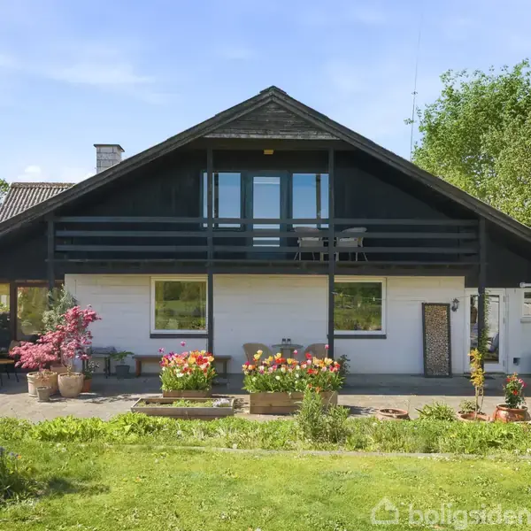 Et hvidt hus med sort tag udført i en klassisk stil står i en velplejet have, omgivet af farverige blomster og grønne træer under en klar blå himmel.