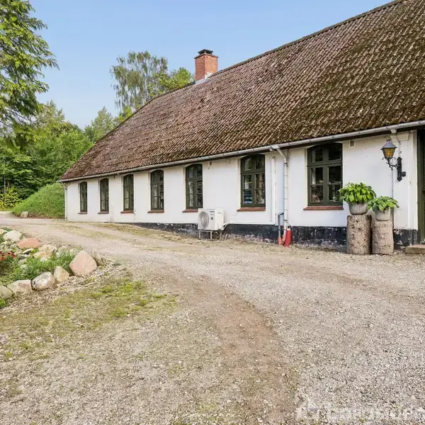 En lang, hvid stuehus med mørkt tegltag står stille i en landlig have med blomsterbede og grusvej, omgivet af høje træer.