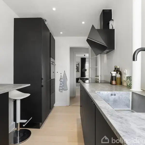 Sort køkkenbord med vask; moderne stil. Højskabe og stol til venstre. Lys træagtigt gulv. Køkkenredskaber ved vask. Vægmonteret emhætte over komfur; lysindfald fra højre.