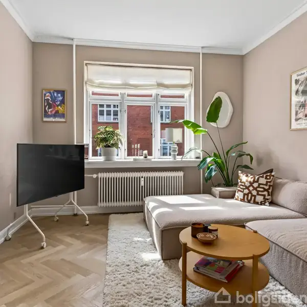 En lys stue med en beige sofa og et lille rundt træbord ved siden af. En TV-skærm står foran vinduet, som har udsigt til en rød murstensbygning. Stueplanter og kunst hænger på væggen.