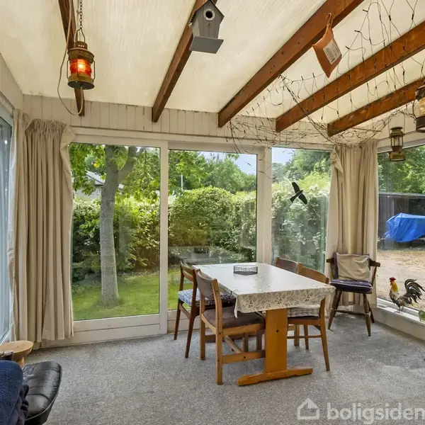 Veranda med bord og stole står stille midt i rummet. Lanterner hænger fra træbjælkerne i loftet. Store vinduer viser en frodig have med grønne træer udenfor.