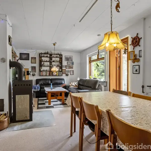 Et spisebord med træstole står i et lyst rum. En brændeovn til venstre, sofaarrangement i baggrunden, reoler og billeder på væggen. Store vinduer giver udsigt til en have.