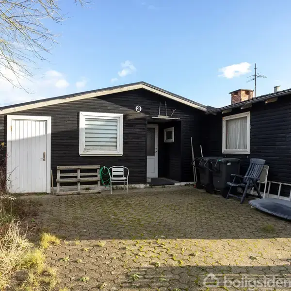 Sortmalet hus med fladt tag, som har træmøbler og affaldsbeholdere placeret ved indgangen. Brostensbelagt gårdsplads og omgivende vegetation, under en klar himmel.