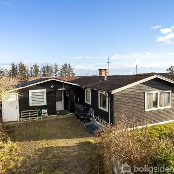 Et sort træhus står stille i en åben, græsklædt have med flisebelagt indkørsel. Omgivelserne inkluderer træer og få huse i baggrunden under en klar blå himmel.