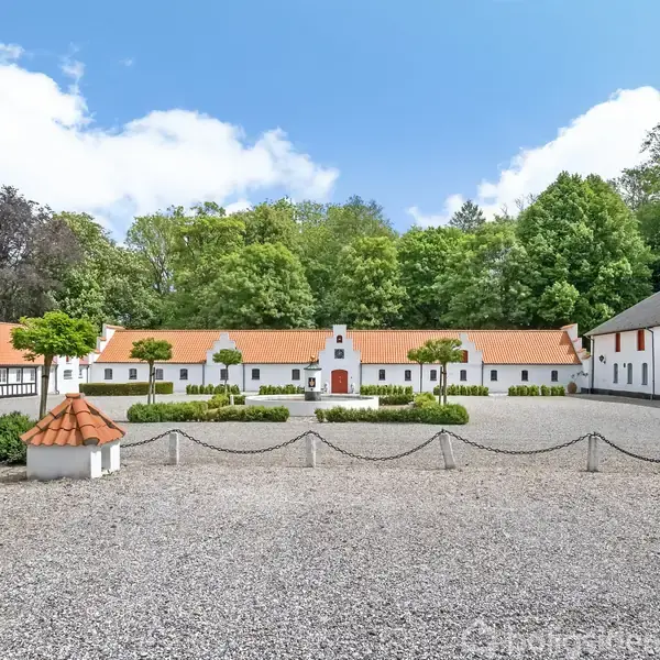En firlænget, hvidkalket gård med røde tegltage. Gårdspladsen er dækket med grus og omkranset af små buske. Bagved er der grønt løv fra høje træer under en blå himmel.