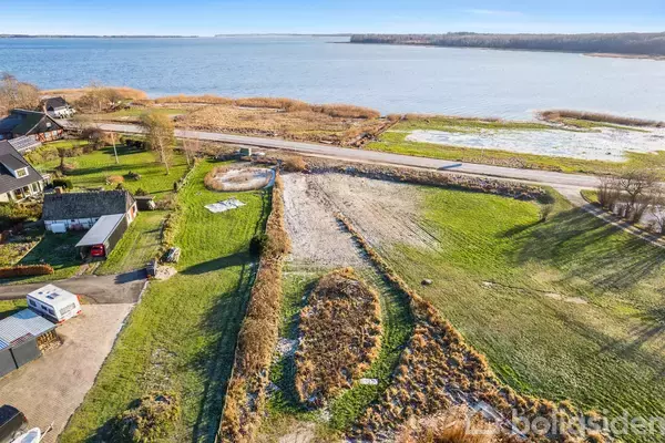 Grønt åbent jordareal strækker sig mod en stille vej, som adskiller det fra en sø i horisonten. Omgivet af huse og biler, ligger arealet i et landligt miljø.