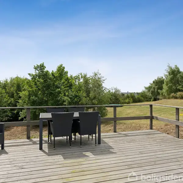 En træterrasse med et bord og fire sorte stole står midt i et frodigt, grønt landskab under en klar blå himmel.