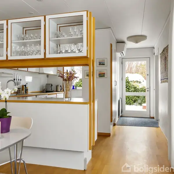 Et moderne køkken har glasvitriner med glas og en hvid bordplade. Bordet i forgrunden har en hvid blomst. I baggrunden ses en gang med en hvid dør.