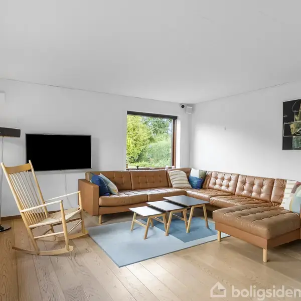 Brun lædersofa i hjørne med farvede puder, ved siden af lænestol og sofabord, på træparketgulv, i lys stue med vindue til grøn udsigt og moderne kunst på væggen.