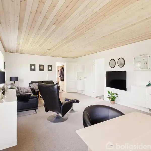 Stue med moderne indretning, hvor sorte lænestole og sofaer er rettet mod et vægmonteret TV. Hvide vægge og lofter i lyst træ. Blomster og vægkunst tilføjer farver.