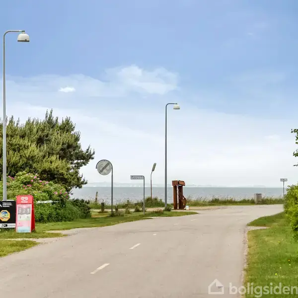 Vej fører mod havet, omgivet af træer og buske. Skilt på venstre side viser teksten "LYSTBÆK". Lamper står langs vejen under en klar blå himmel.