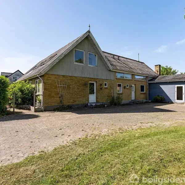 Et gult murstenshus med skråt tag står stille omgivet af grønt græs og træer under en klar blå himmel.