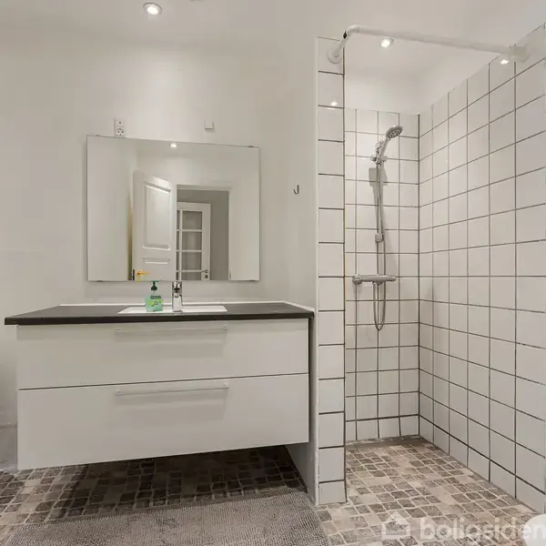 Badeværelse med hvidt skab og vask under et spejl. Til højre er en brusekabine med hvide fliser. Der er en kurv på gulvet og lys grå flisebelægning.