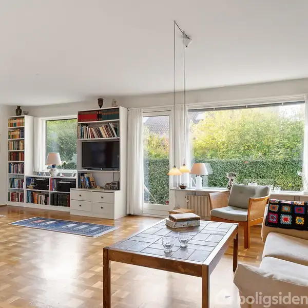 Stue med sofa og sofabord, omgivet af store vinduer med udsigt til en have. Rummet har trægulv, reoler fyldt med bøger og en skænk med fjernsyn.