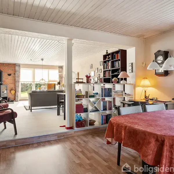En spisestue med rødt dugklædt bord fører til en lys stue. Baggrund indeholder reoler, en sofa, brændeovn, og store vinduer med udsigt til haven.