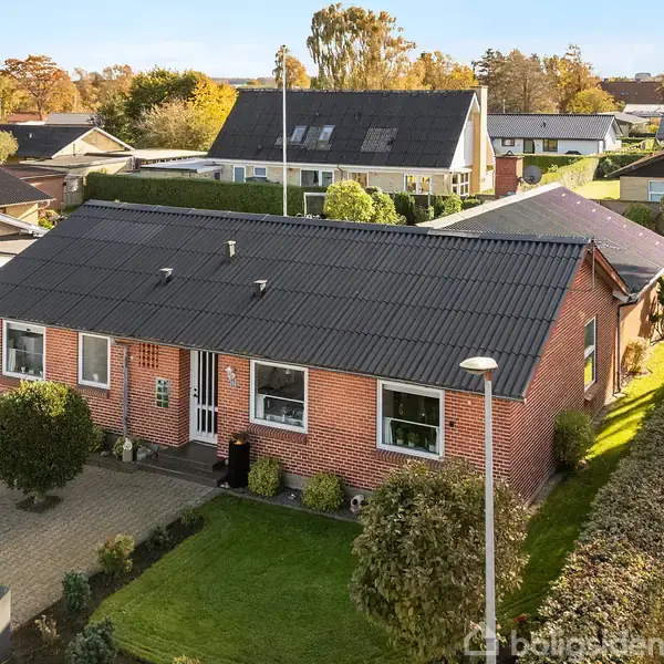 En etplans murstensvilla med sort tag står foran en indkørsel og velplejet have. Omkringliggende hække og træer skaber en rolig forstadsmiljø. En rød bil ses i carporten.