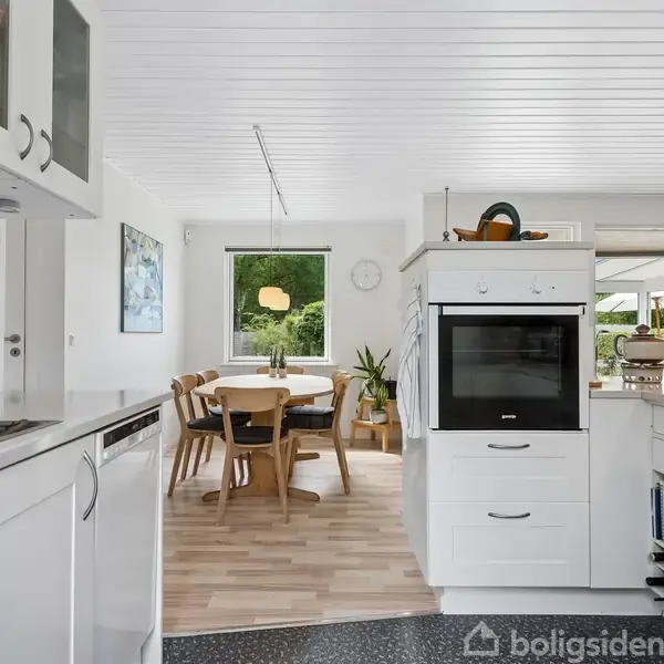 Et moderne køkken med hvide skabe og indbyggede apparater leder mod en spisestue med et rundt træbord og stole. Store vinduer giver udsigt til en grøn have.