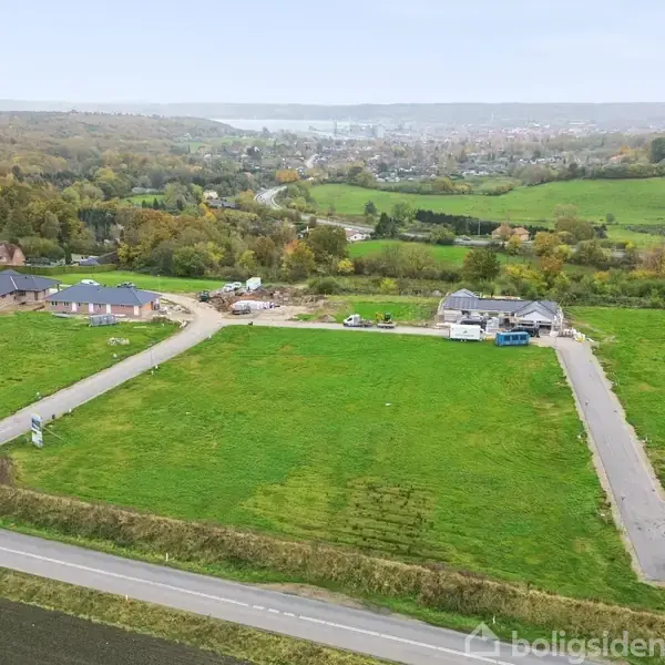 Grøn mark omgivet af veje og bygninger under konstruktion i bakkelandskab, med en by i baggrunden.