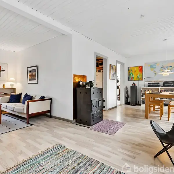 Et moderne stueområde med lys indretning. Sofa og stole står tæt på et vindue, mens et spisebord med stole er i baggrunden ved siden af et klaver. Kunst hænger på væggene.