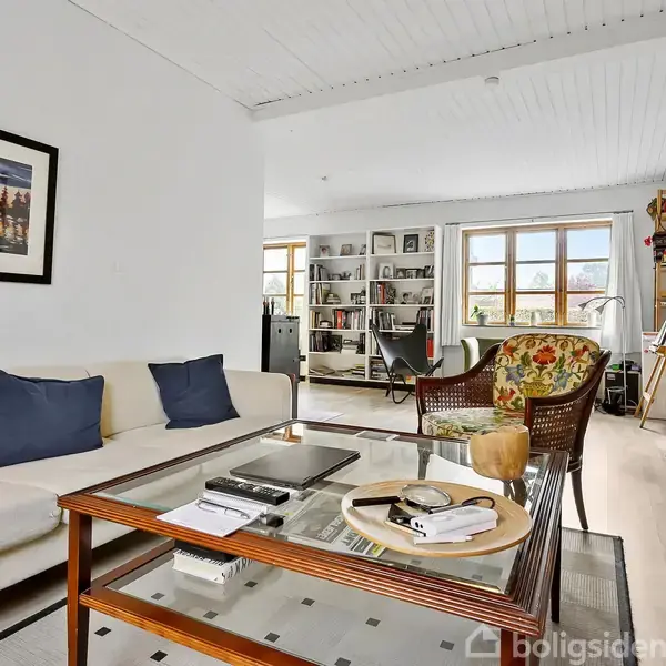 En lys stue med en hvid sofa og blå puder ved siden af et træsofabor med bøger. Væggene prydes med billeder. Baggrunden viser reoler og store vinduer.