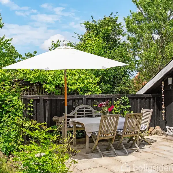En terrasse med et bord og seks stole står under en stor, hvid parasol. Det omkringliggende område er grønt og frodigt med træer og en sortmalet træbygning.