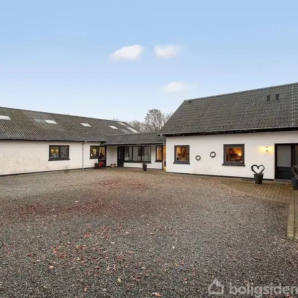 En hvid, vinkelformet ejendom står stille på en grusbelagt gårdsplads, omgivet af bartræer og en klar blå himmel. Der er flere vinduer og en hoveddør med planter.