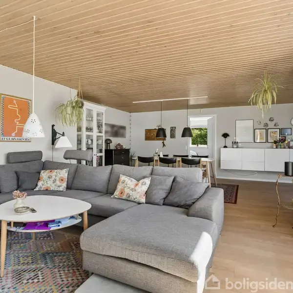 Grå sofa med farverige puder står i en moderne stue med trægulv og træloft. Over sofaen hænger plakater. Rummet har spisebord, planter og store vinduer til højre.