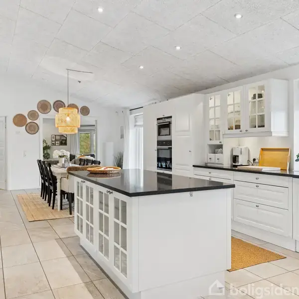 Et moderne køkken med en stor central køkkenø i sort og hvid. Køkkenet er omgivet af hvidt skabsinventar og store vinduer, der bringer naturligt lys ind.