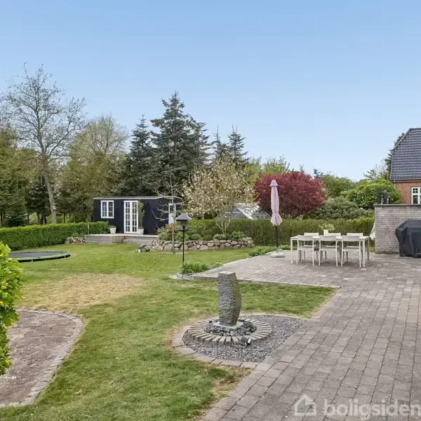 En have med flisebelagt sti fører til et spisebord med parasol tæt på et hus. Bagerst ses en sort skur, græsplæne og forskellige træer og buske.