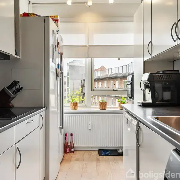En moderne køkkenindretning med hvide skabe og sorte bordplader. Kaffemaskine står ved vasken, knive i en holder ved siden af. Et stort vindue viser byudsigt med bygninger udenfor.