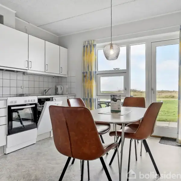 Et køkken har hvide skabe, komfur og spisebord med fire brune stole. Store vinduer med farverige gardiner giver udsigt til grønt landskab udenfor. En moderne lampe hænger over bordet.