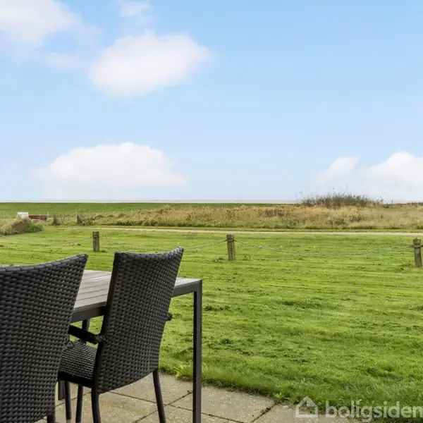 Et havebord med kurvestole står på en flisebelagt terrasse, med udsigt til en velplejet græsplæne og en åben himmel i baggrunden.