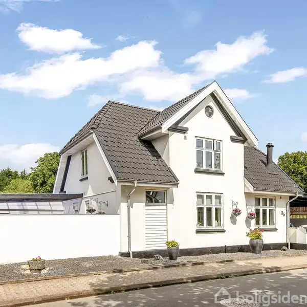 Hvidt hus med gråt tegltag står stille ved en asfalteret vej. Omgivet af grønne træer og buske, med en garage til højre. Blå himmel med spredte skyer ovenover.