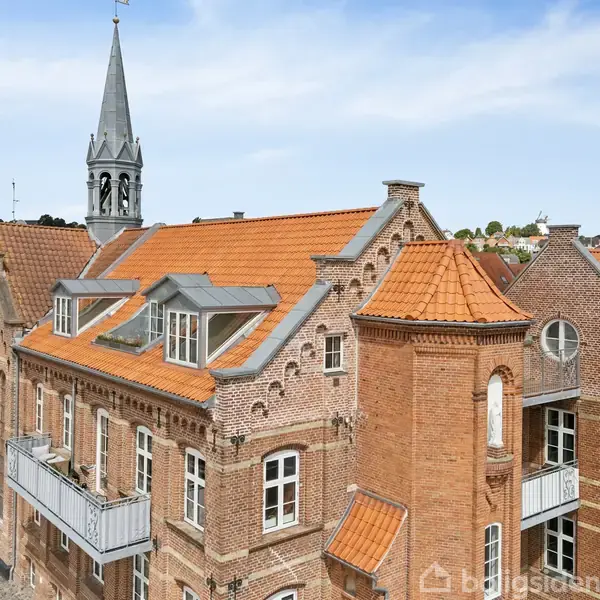 Bygning med røde mursten og orange tegltag har flere altaner og vinduer. I baggrunden ses kirketårn og bylandskab under blå himmel.
