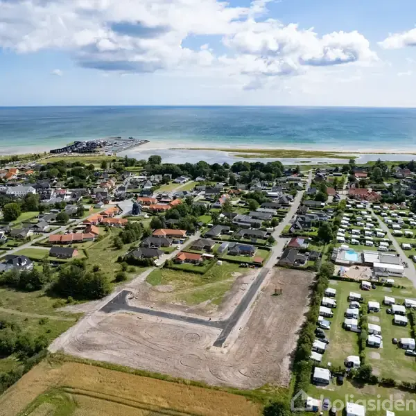 Luftfoto viser et kystnært byområde med huse, campingplads og åbent landskab. Havet strækker sig i baggrunden under en delvist skyet himmel, mens jorden i forgrunden er forberedt til byggeri.