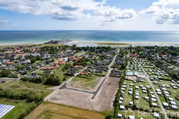 Et landskab med huse og campingplads beliggende ved kysten. Byen udstrækker sig mod havet i baggrunden under en blå himmel med skyer. Fjern lysning afslører en strandpromenade.