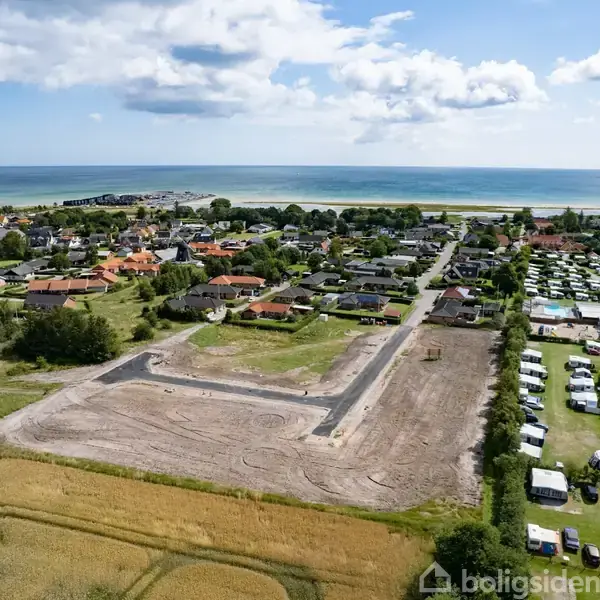 Luftfoto af en grund under udvikling omgivet af marker, huse og en campingplads. I baggrunden ses en kystlinje med havet og en blå himmel med skyer.