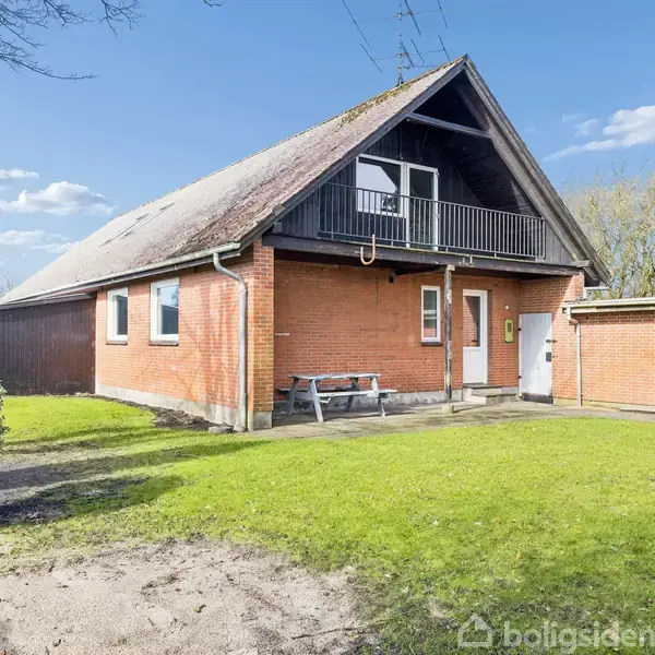 Rødt murstenshus med skråt tag og altan. En bænk står ved hoveddøren. Huset ligger på en grøn græsplæne omgivet af buske, under en klar blå himmel med skyer.