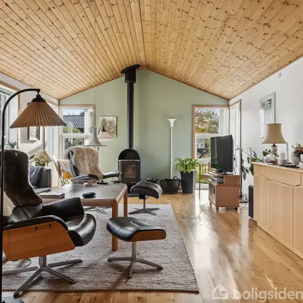 Stue med lænestol og fodskammel, placeret på gulvtæppe. I baggrunden ses en brændeovn. Rummet har trægulv og træloft samt vinduer med udsigt til natur.