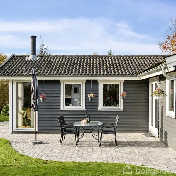 Gråt træhus står stille, omgivet af grøn have. Foran huset står et rundt bord med sorte stole og en parasol. Blomster hænger under vinduerne, omkranset af træer.
