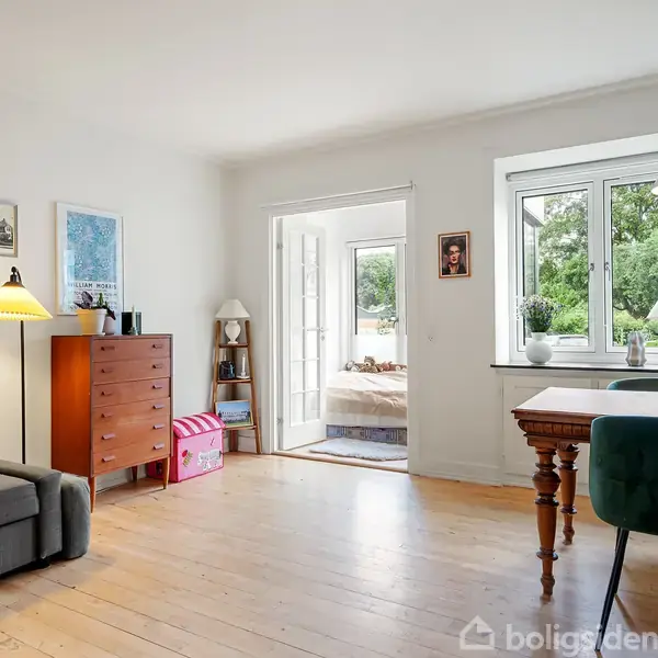 En stue med en grå sofa til venstre, en kommode, et lysende gulvlampe og en åben dør til et soveværelse. Til højre står et træbord med blomster og to stole.