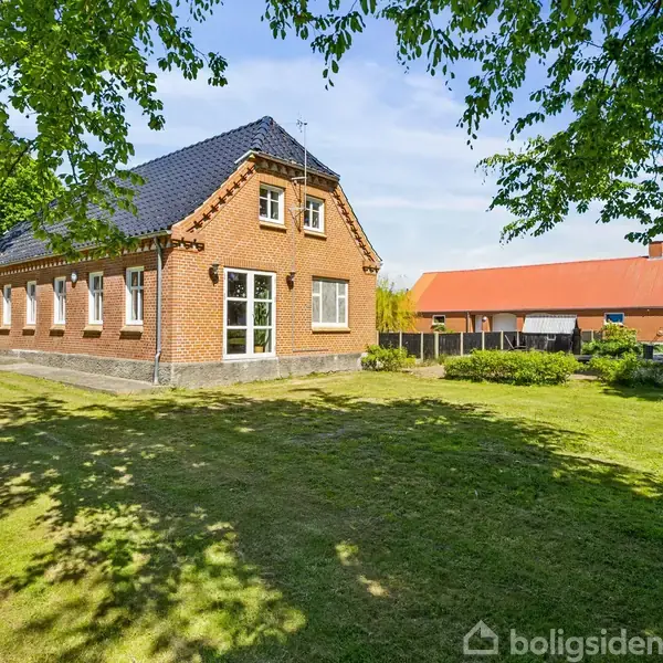 En rød murstensbygning med mørkt tag står på en velplejet græsplæne. Der er træer i forgrunden og en anden bygning med rød tag i baggrunden under en klar blå himmel.