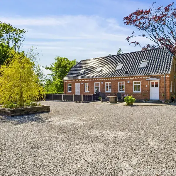 En rød murstensbygning står stille på en grusplads omgivet af træer. En lille, træbeplantet have ses foran huset.