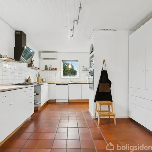 Et moderne køkken med hvide skabe og skuffer strækker sig langs væggene. En skammel med en sort forklæde hænger ved siden af. Rummet har røde klinkegulve og et vindue med udsigt.